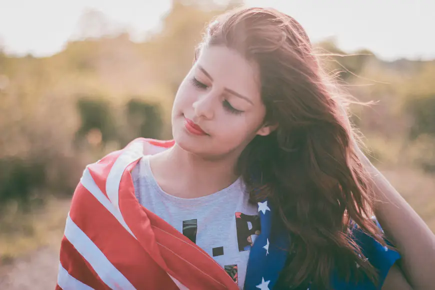Outdoor headshot of a woman with long brown hair wrapped in an American flag, eyes closed in warm golden light